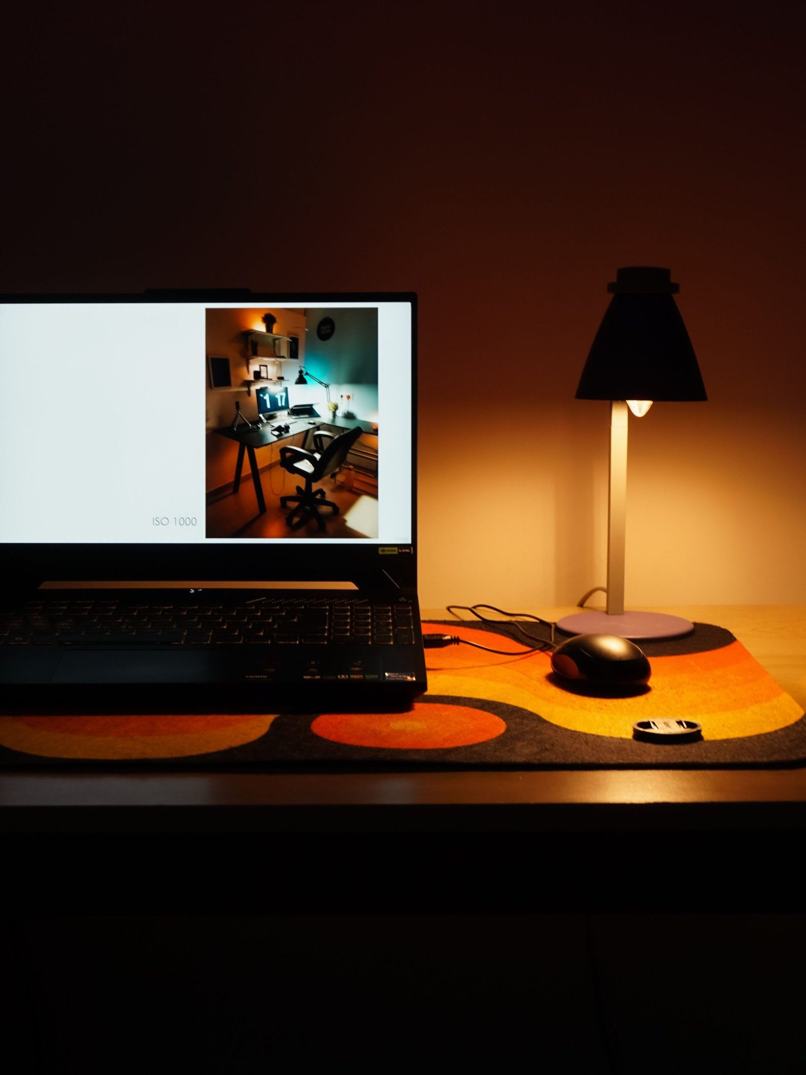 A dimly lit desk with a laptop, mouse, and lamp creating a warm workspace ambiance.